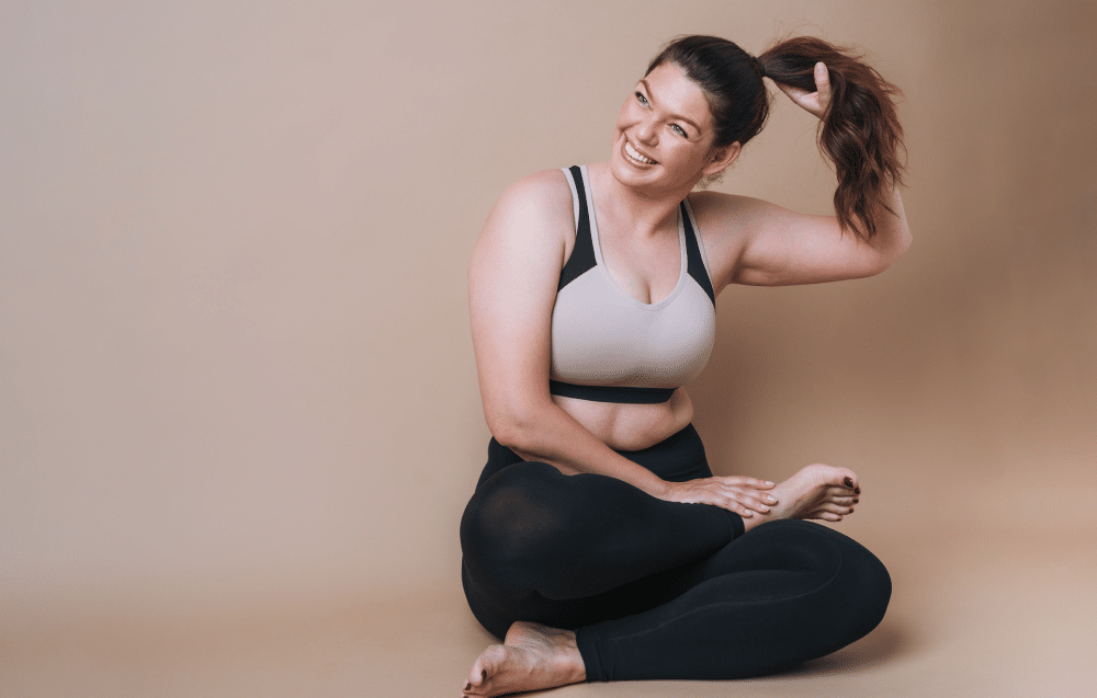 A woman in exercise attire sits cross-legged on the floor, smiling radiantly as she adjusts her ponytail with one hand, embodying the vitality and renewal that REGENICS promotes.